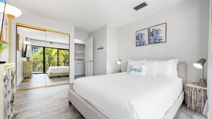 Une chambre moderne avec un grand lit, une table de chevet, une commode et une vue sur la cuisine et le balcon.