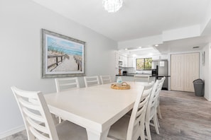 Un coin repas avec une table et des chaises blanches, un tableau représentant une scène de plage et, au fond, une cuisine.
