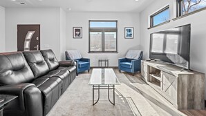 Ein modernes Wohnzimmer mit einem Ledersofa, zwei blauen Sesseln, einem Glastisch, einem Fernseher auf einer Holzkommode und einem großen Fenster.