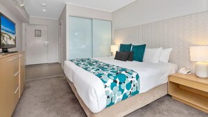 Moderni hotellihuone, jossa on suuri sänky, televisio, vaatekaappi ja työpöytä, jossa on lamppu.
