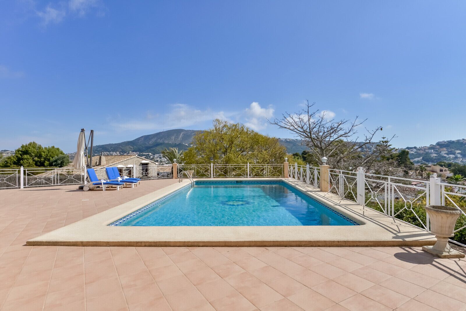 A rectangular swimming pool with a white tiled edge, surrounded by a white fence. There are two blue lounge chairs and a single umbrella on the poolside. The pool is set on a tiled deck, and the background features a mountainous landscape under a clear blue sky.
