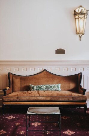 Un canapé marron agrémenté d’un coussin à motifs floraux, une petite table et un luminaire fixé au mur.