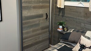 Una habitación con puerta corredera, cama con ropa de cama estampada, mesita de noche con una planta y un reloj, y ventana con vista a árboles.