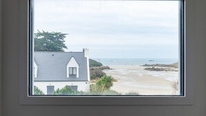 Une vue sur la côte, aperçue à travers une fenêtre, comprenant une maison avec un toit gris et des murs blancs, une plage de sable et la mer.