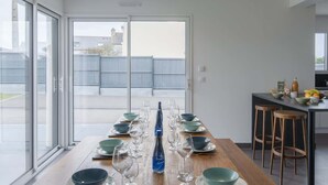 Une longue table à manger, dressée pour un repas, avec de la vaisselle et des bols, dans une cuisine moderne dotée de grandes fenêtres et offrant une vue sur la ville.