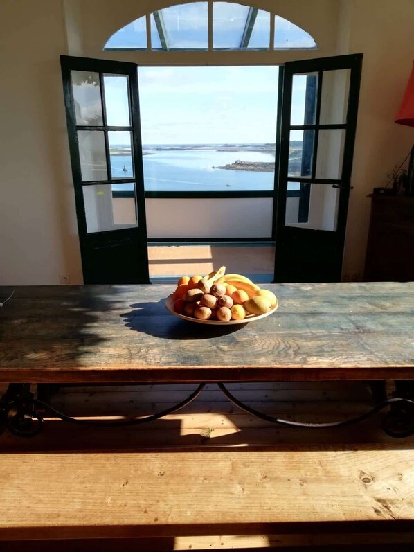 Una mesa con un bol de fruta en una habitación con vistas al mar a través de puertas abiertas.