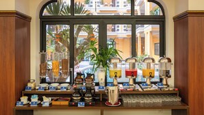 A breakfast bar with various food and drink dispensers, a coffee station, and a view of the outside buildings through a large arched window. - Soleil by Soluna - A Charming Touch of Hoi An's Heritage (Da Nang)