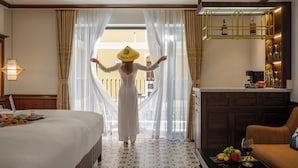 A hotel room with a bed, a dining table with fruits and a glass of wine, a sofa, and a minibar with bottles and glasses. - Soleil by Soluna - A Charming Touch of Hoi An's Heritage (Da Nang)