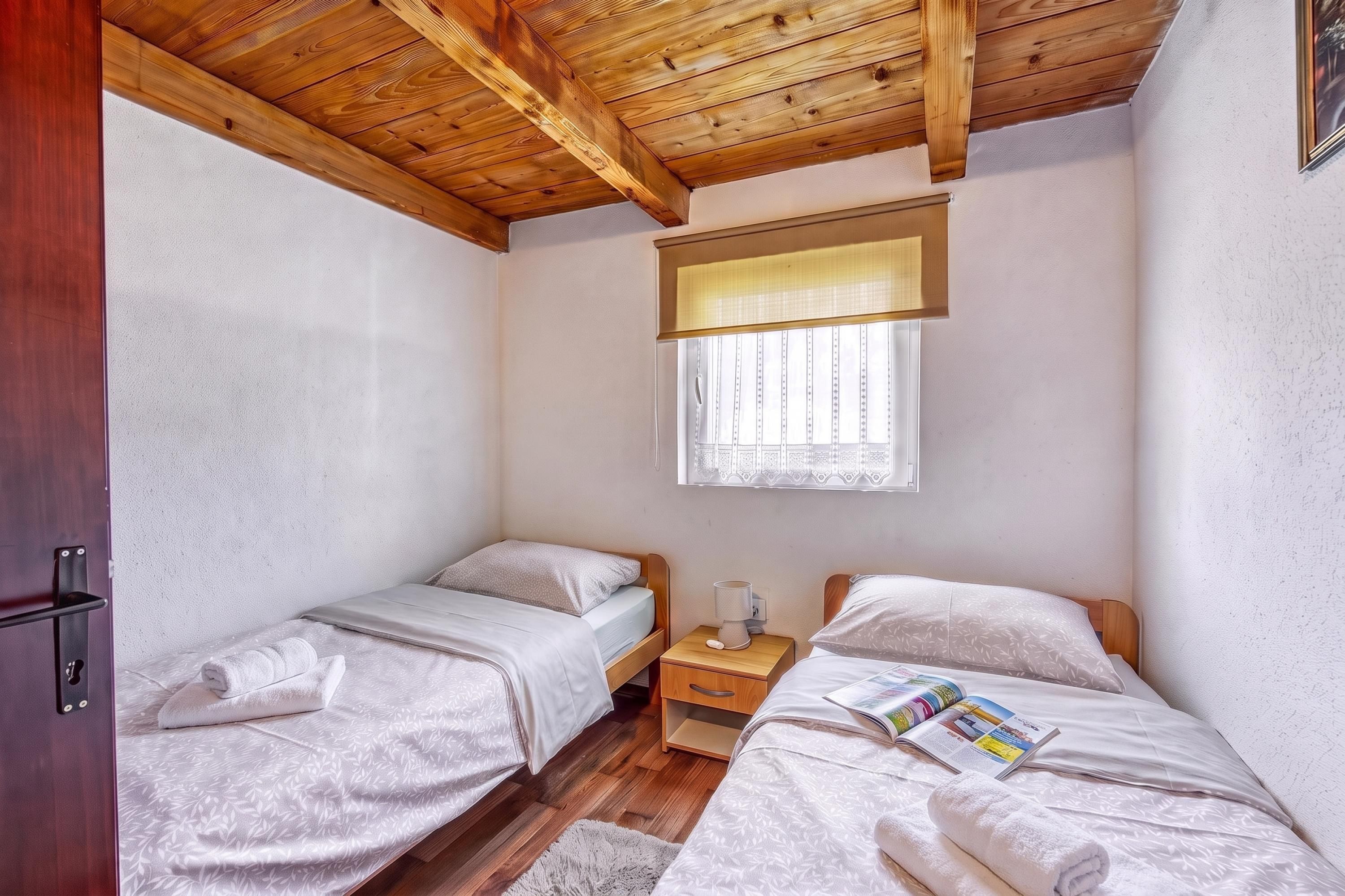 Een knus slaapkamer met twee eenpersoonsbedden, een houten plafond, een raam met zonwering en een nachtkastje met een lamp.