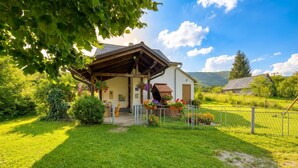 A small house with a wooden porch, surrounded by a well-maintained lawn and a fenced garden area.