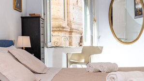 A hotel room with a bed, a desk, a chair, a wardrobe, a mirror, and a window with a view of a building.
