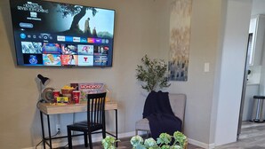 Una sala de estar con una mesa de centro de madera, un televisor mostrando la interfaz de un servicio de streaming, una zona de comedor con sillas y una mesa, y una planta decorativa.