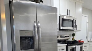 Un refrigerador de acero inoxidable con dispensador de agua, un microondas y un horno en una cocina moderna con armarios blancos y piso de madera.