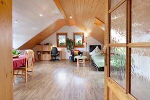 Ein Zimmer mit schrägverdeckter Decke, Holzboden, einem Esstisch mit Stühlen, einem kleinen runden Tisch, einem Bett, einem Schreibtisch mit Stuhl und zwei Fenstern mit Vorhängen.
