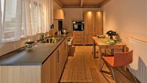 A compact kitchen with wooden cabinets, a built-in oven, a dining area with a table and chairs, and a window with sheer curtains. - Holiday Apartment 'Albfewo' with Terrace, Garden and EV Charging Station (St. Johann)