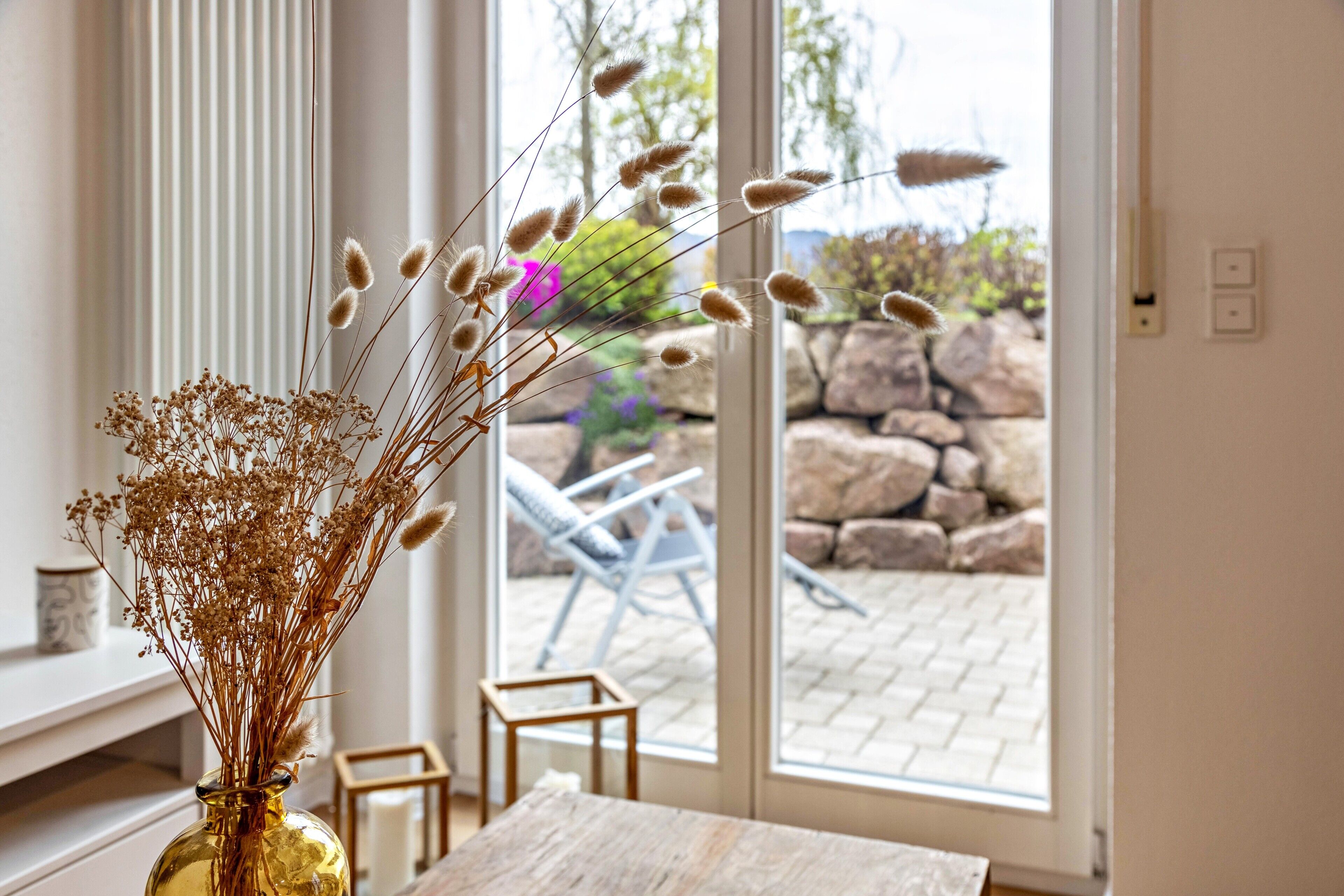A room with a large window, a vase with dried plants, a wooden table, and a chair.