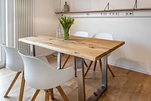 Une table de salle à manger en bois, accompagnée de quatre chaises blanches, un vase rempli de fleurs jaunes et un luminaire moderne fixé au mur.
