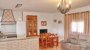 Een woonkamer met een bank, eettafel en stoelen. Er is een keuken met een bakstenen aanrecht en kastjes. Een kroonluchter hangt aan het plafond, en gordijnen bedekken de ramen.