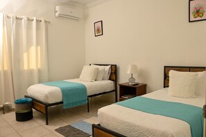 A bedroom with two beds, a small stool, a nightstand, and a window with curtains. - Villa Serenity by the water (Providenciales)