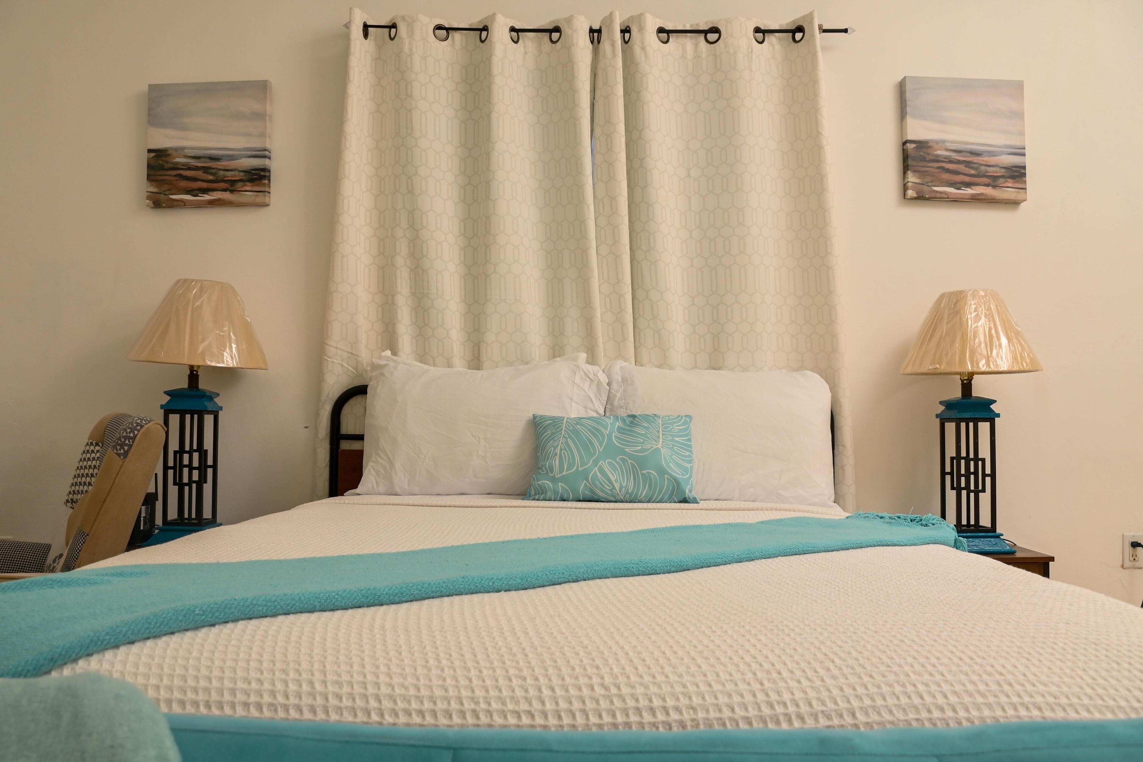 A bedroom with a bed, two wall-mounted lamps, a window with a curtain, and two paintings on the wall.