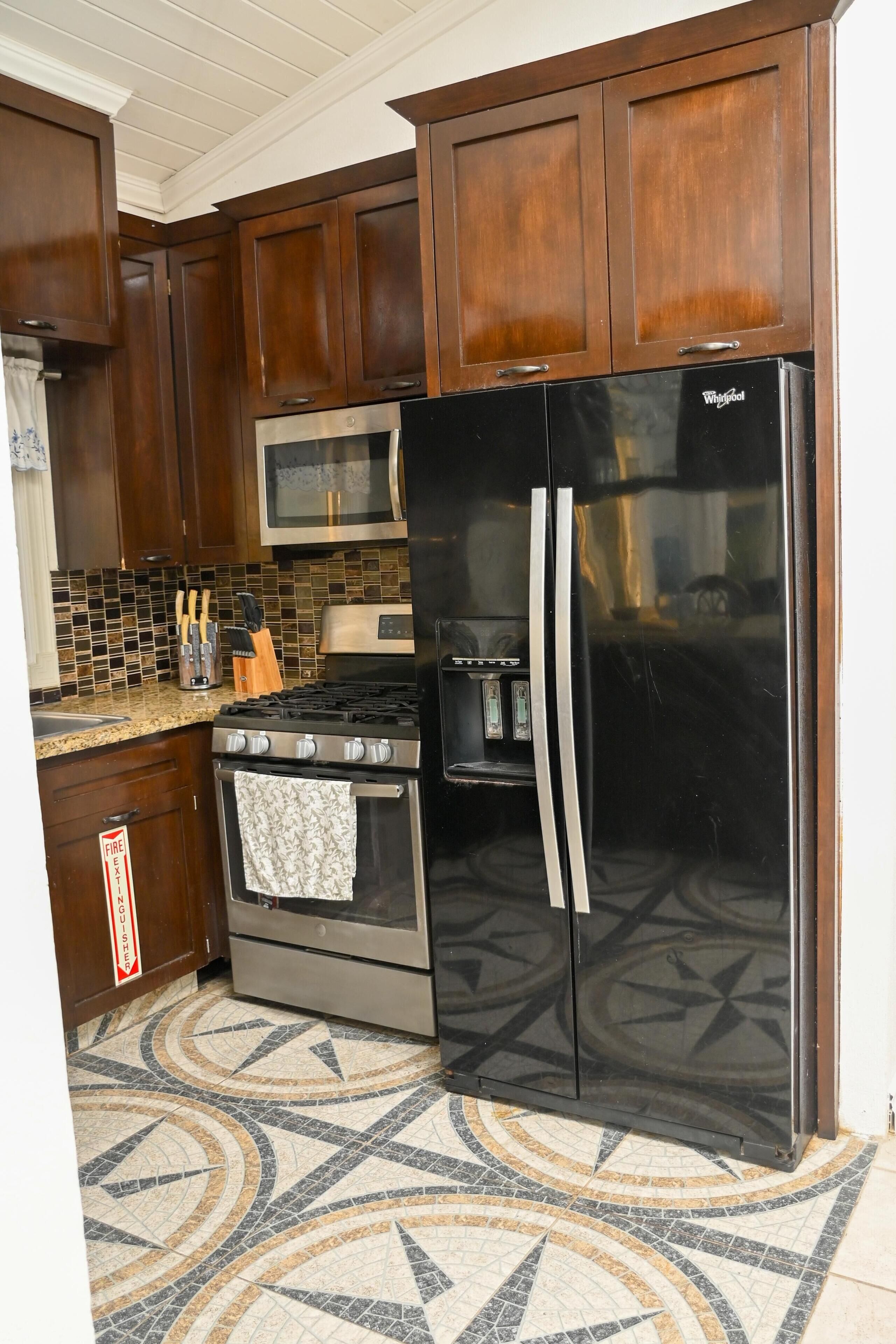 A kitchen with dark wooden cabinets, a stainless steel refrigerator, a microwave, a gas stove, and a patterned tile floor.