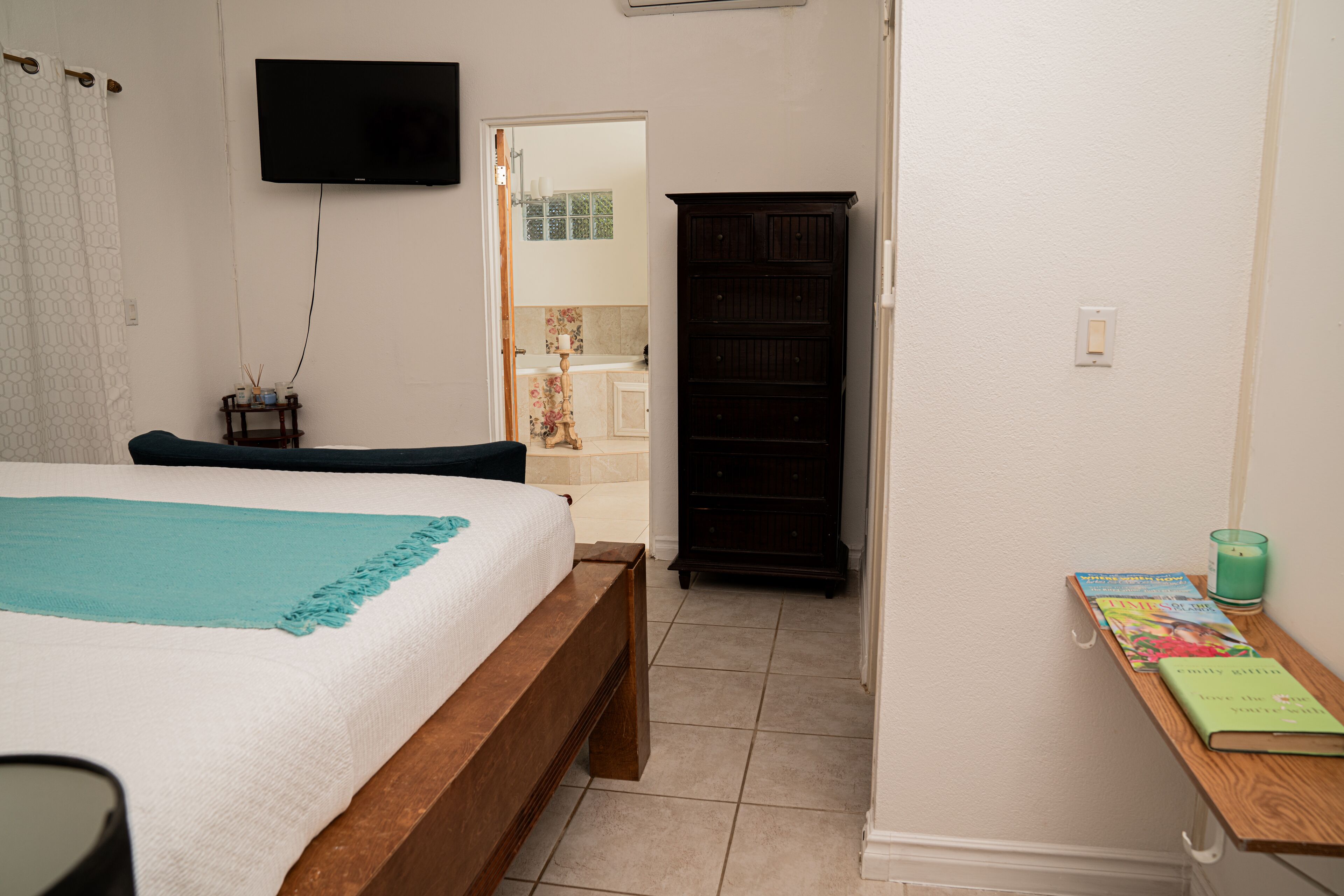 A bedroom with a bed, a TV mounted on the wall, a wooden cabinet, and a small table with books and a candle.