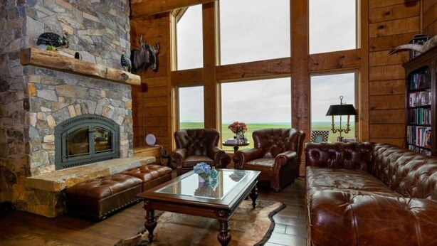 Un interior rústico con chimenea de piedra, mobiliario de cuero y amplias ventanas con vistas a un campo.
