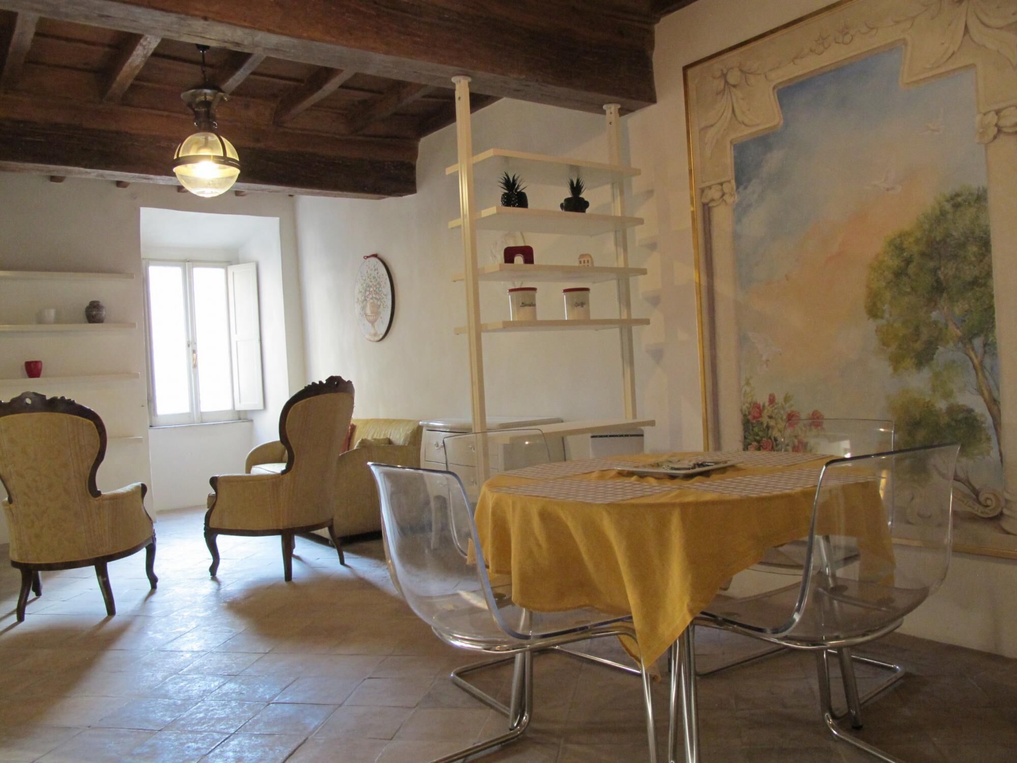 A dining area with a table covered by a yellow tablecloth, surrounded by clear chairs. There are two wooden chairs with curved backs, a white shelf with decorative items, and a painting of a tree on the wall.
