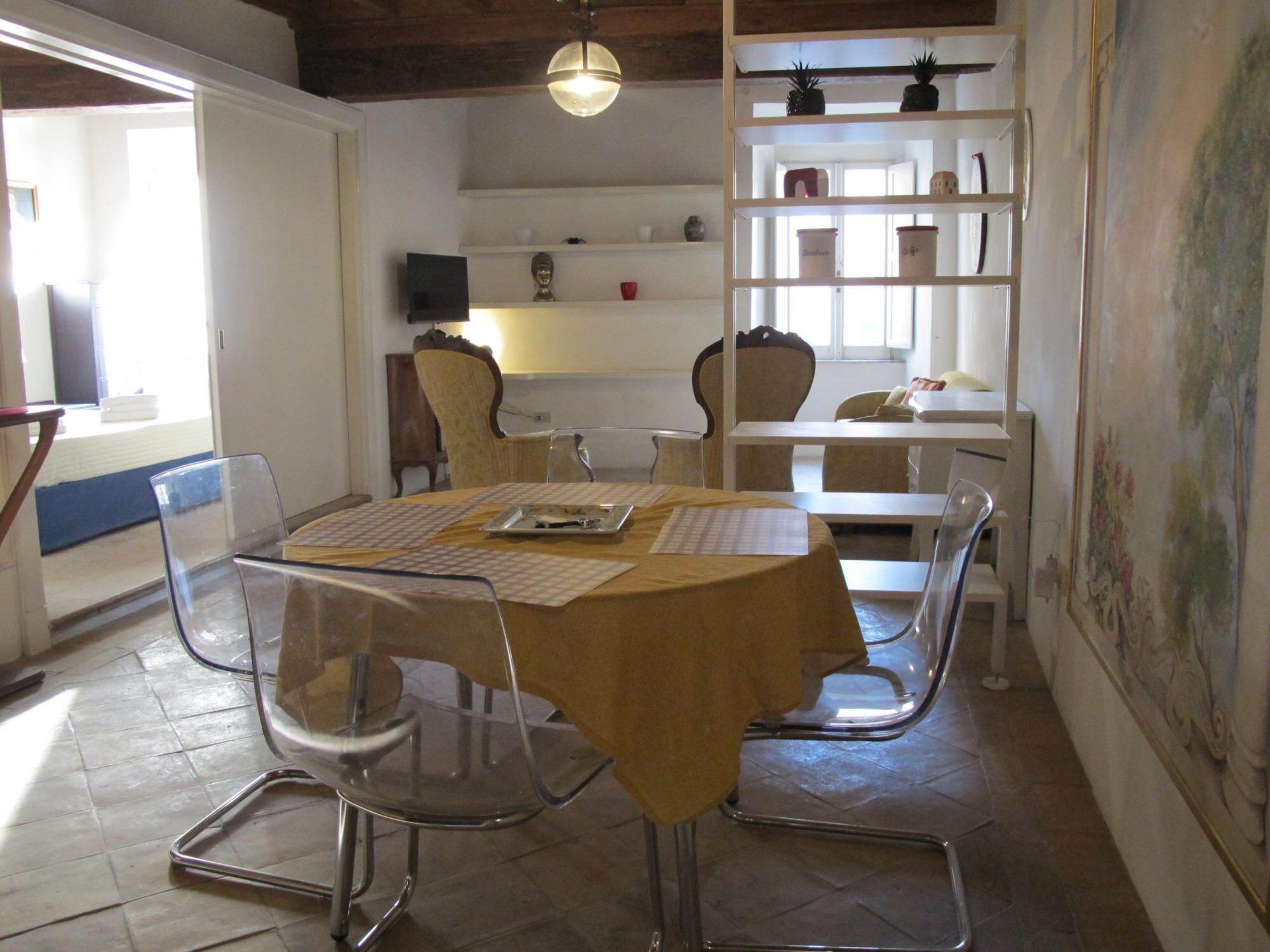 A dining area with a round table, clear chairs, and a yellow tablecloth. There is a shelf with decorative items and a painting on the wall.