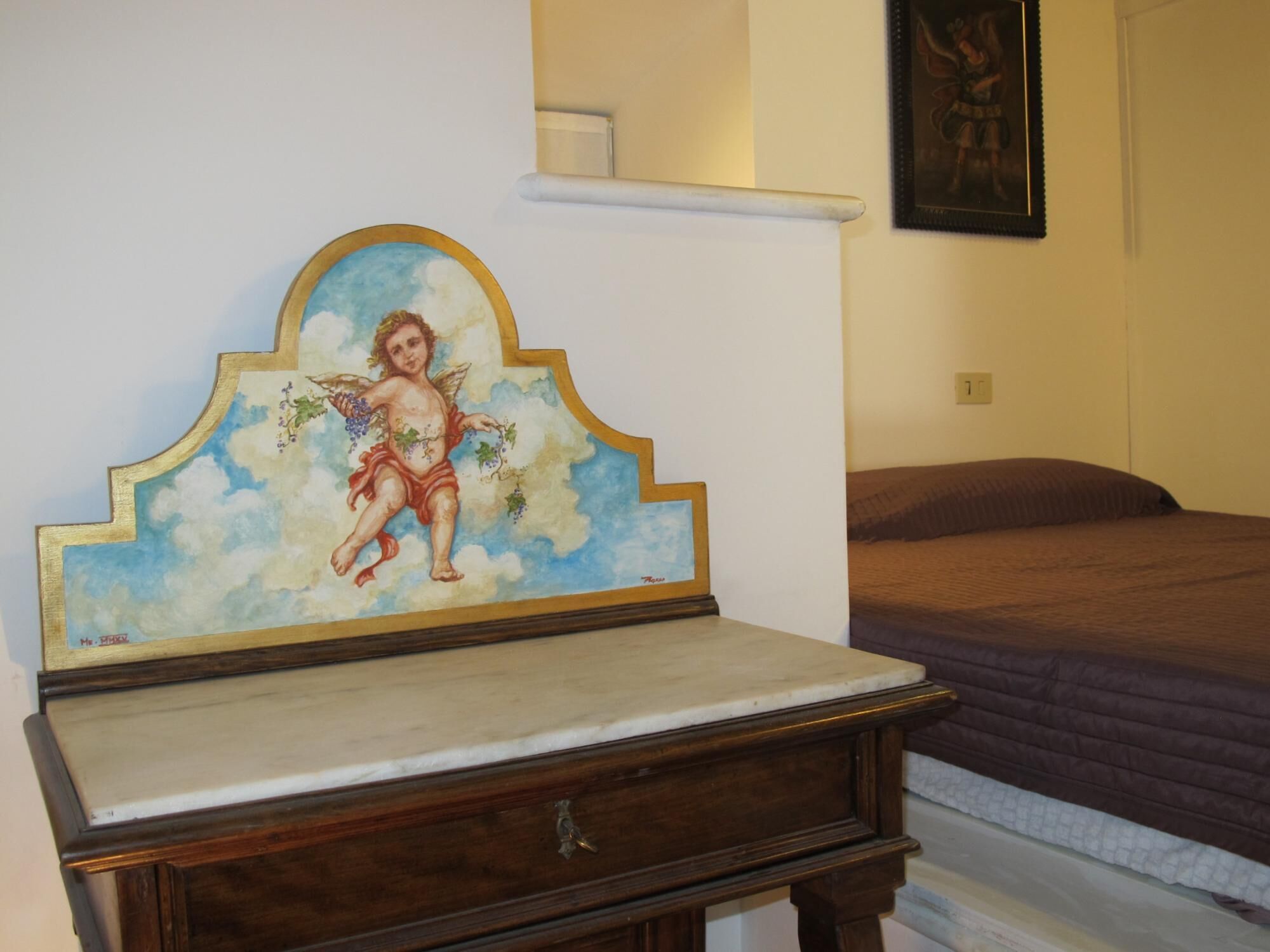 A room with a bed, a small wooden table with a marble top, and a wall-mounted shelf with a framed picture.