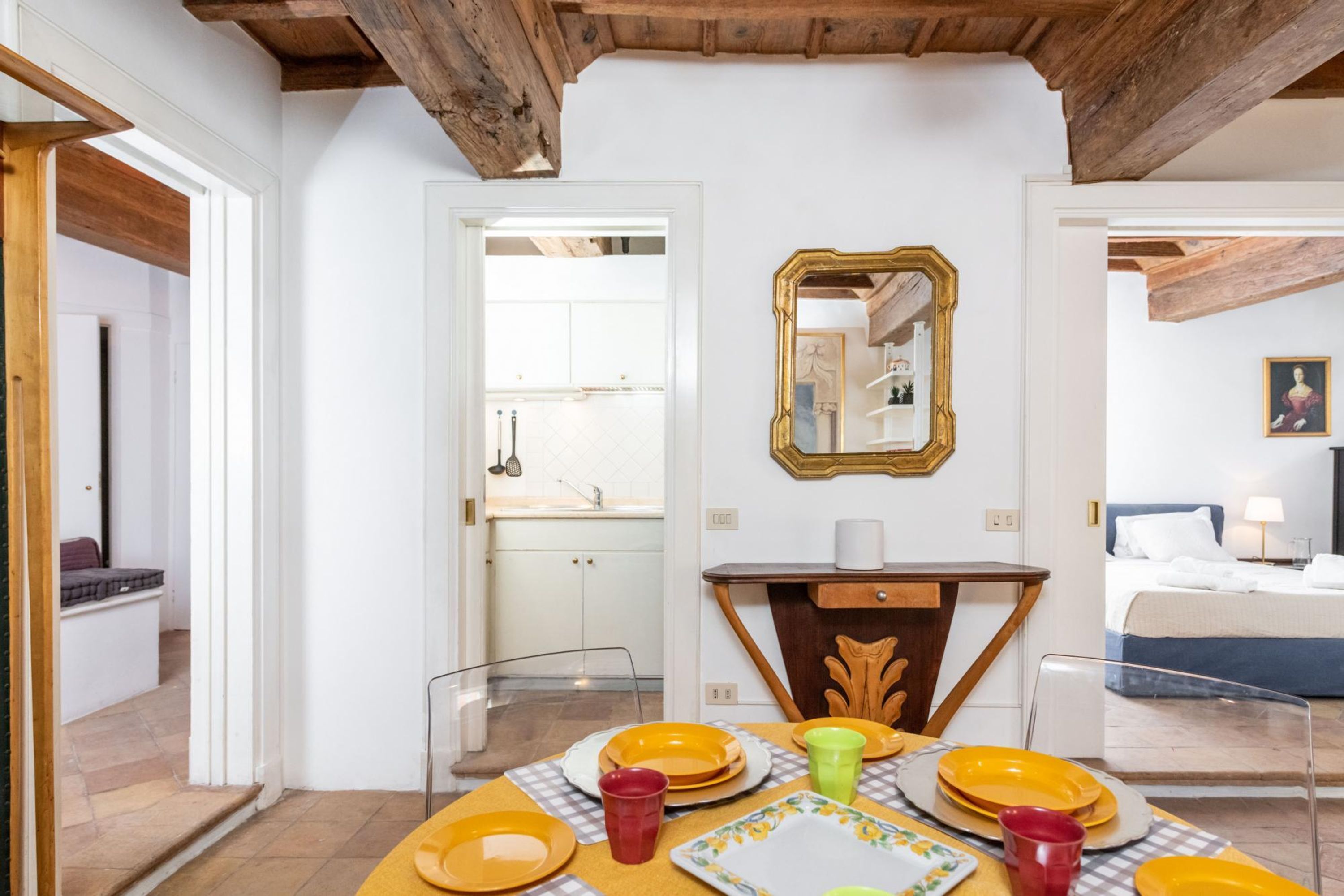 A dining area with a table set for four, a mirror above the table, and a bed in the adjacent room.