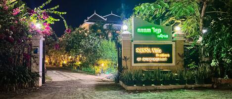 A well-lit entrance with a sign for Sanna Villa Resort & Spa, surrounded by lush greenery and flowering plants.