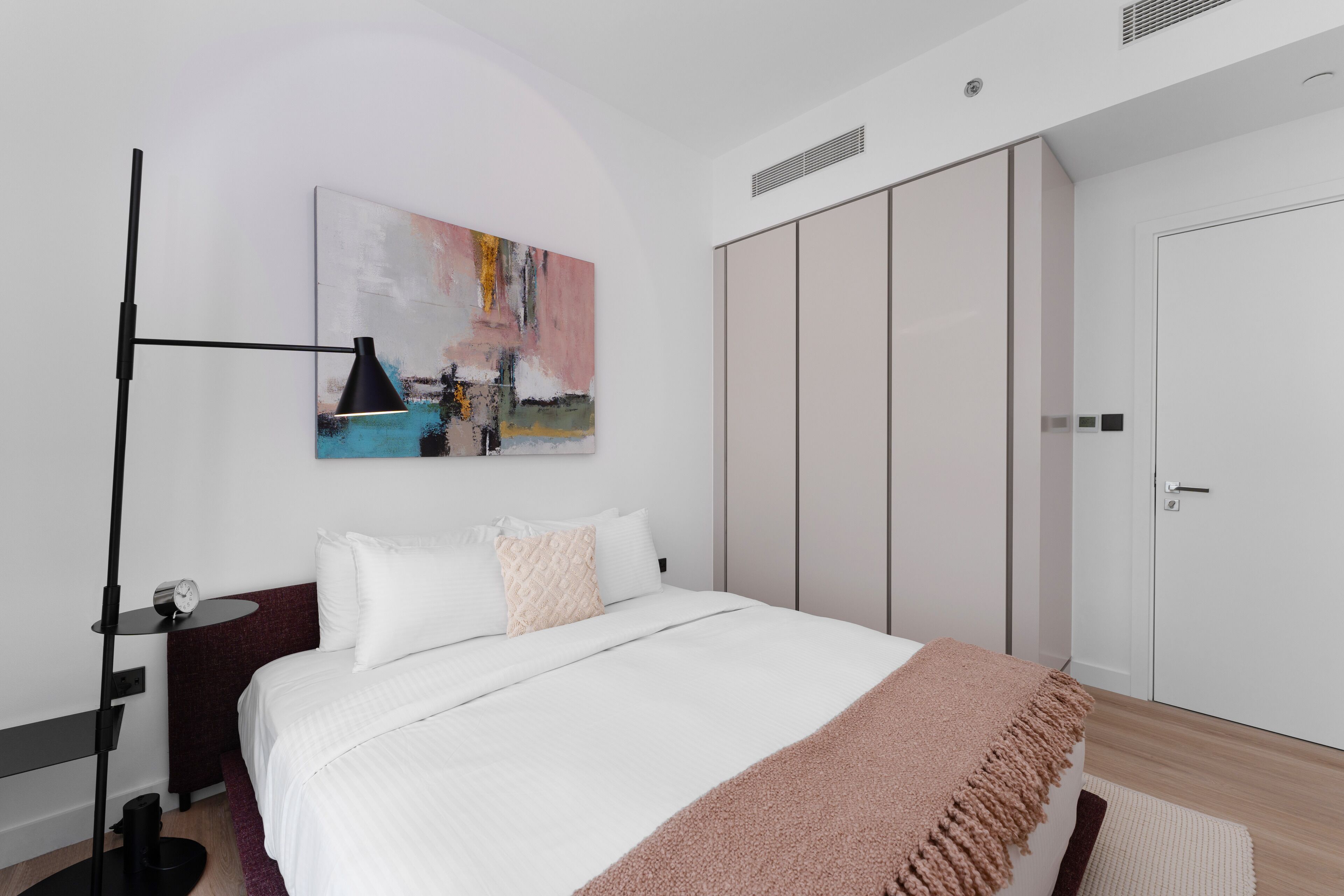 A bedroom with a bed, a bedside table with a lamp, a wall-mounted painting, and a wardrobe.