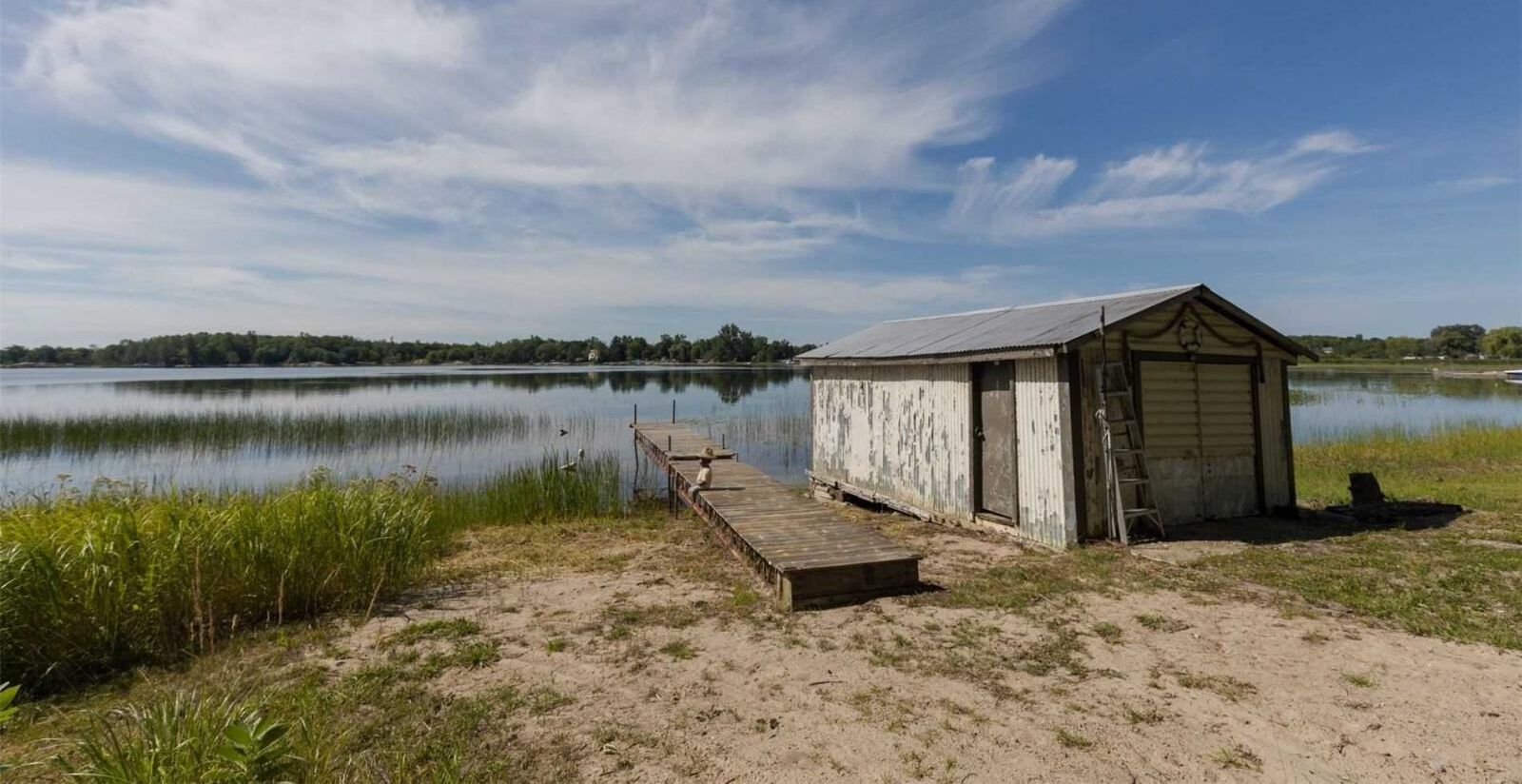 A small, weathered wooden structure with a metal roof by a calm lake, a wooden dock leading to it, and a ladder beside the structure.