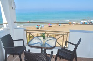 A balcony with wicker chairs and a glass table, overlooking a beach with umbrellas and people.