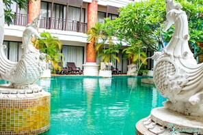 A pool area with two white seahorse statues, a tiled pool edge, and a building with balconies and potted plants in the background.