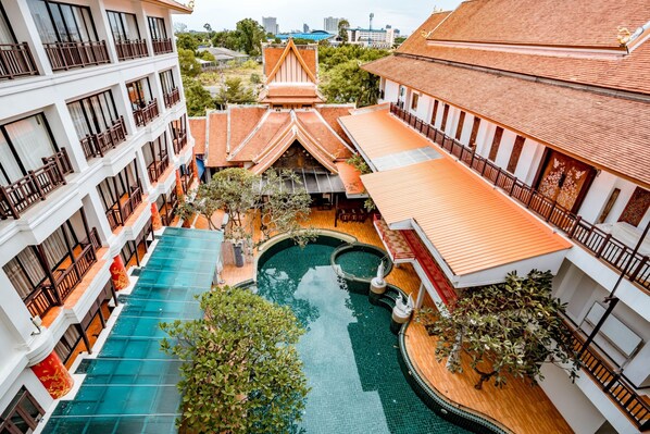 A multi-story hotel with a swimming pool, a courtyard with a traditional roof, and a view of the surrounding cityscape.