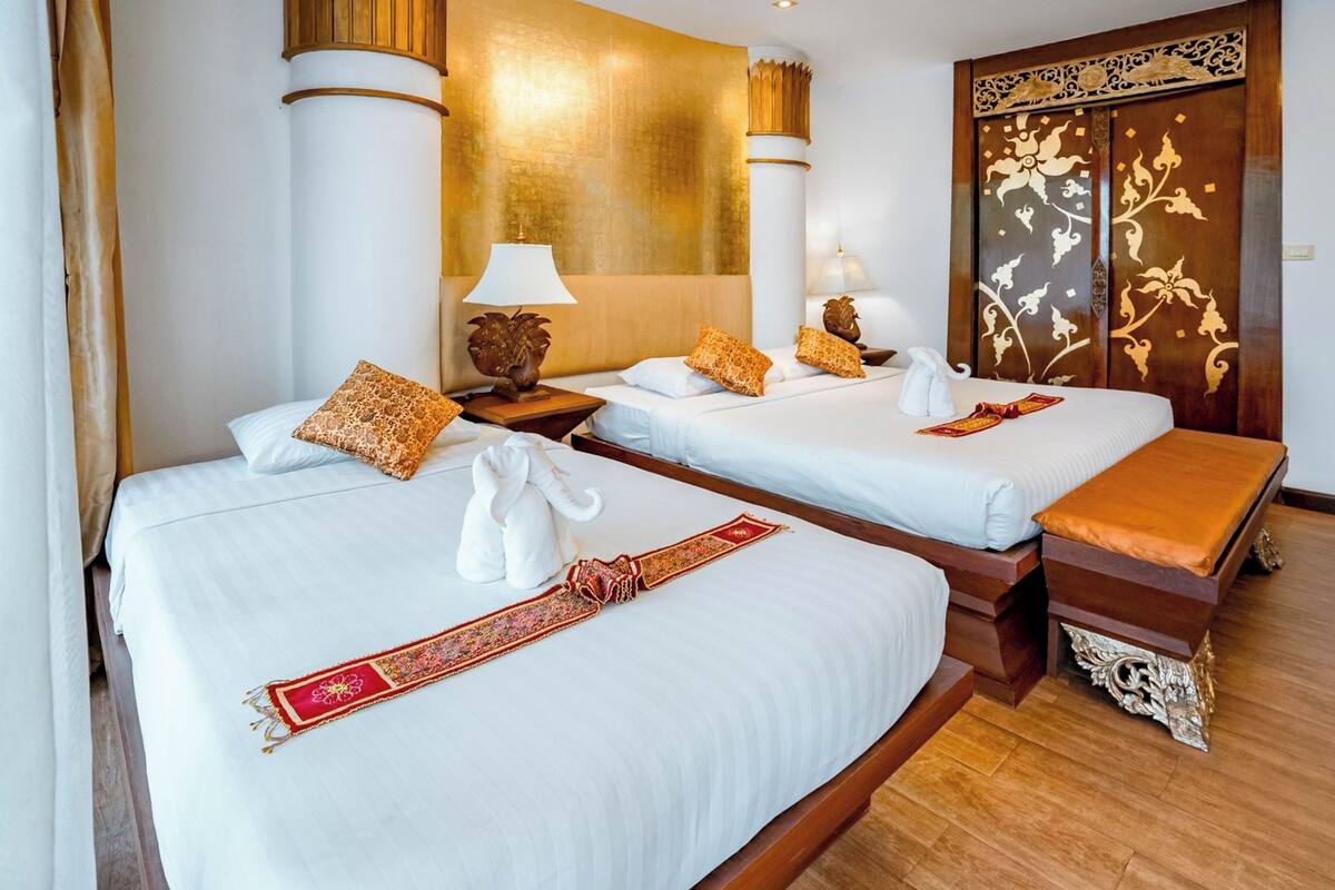 Habitación de hotel con dos camas, cada una con ropa de cama blanca y almohadas decorativas. Hay dos mesitas de noche con lámparas, un banco de madera y un panel decorativo en la pared.