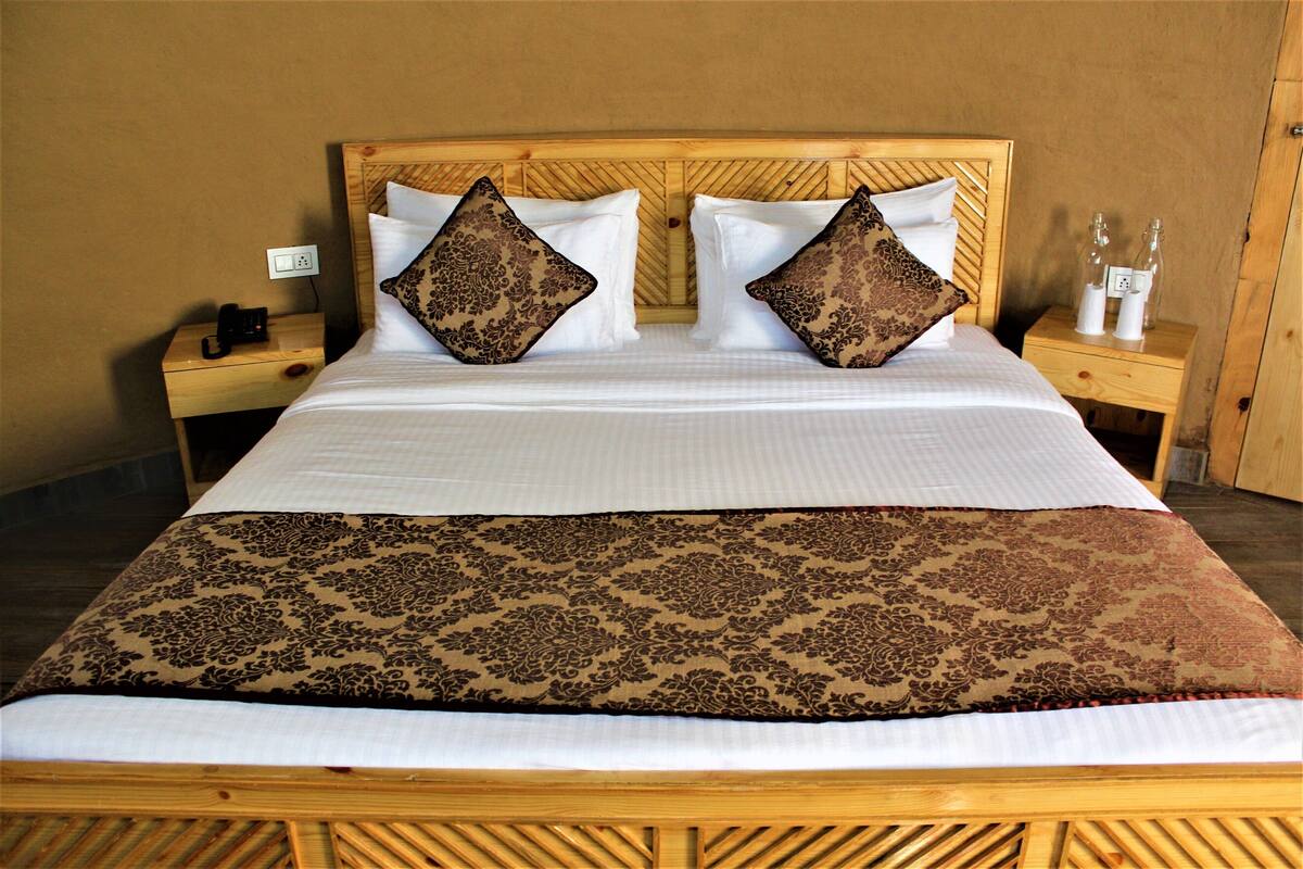 A bed with a patterned bedspread, two decorative pillows, a wooden headboard, and a nightstand with a phone and a bottle.