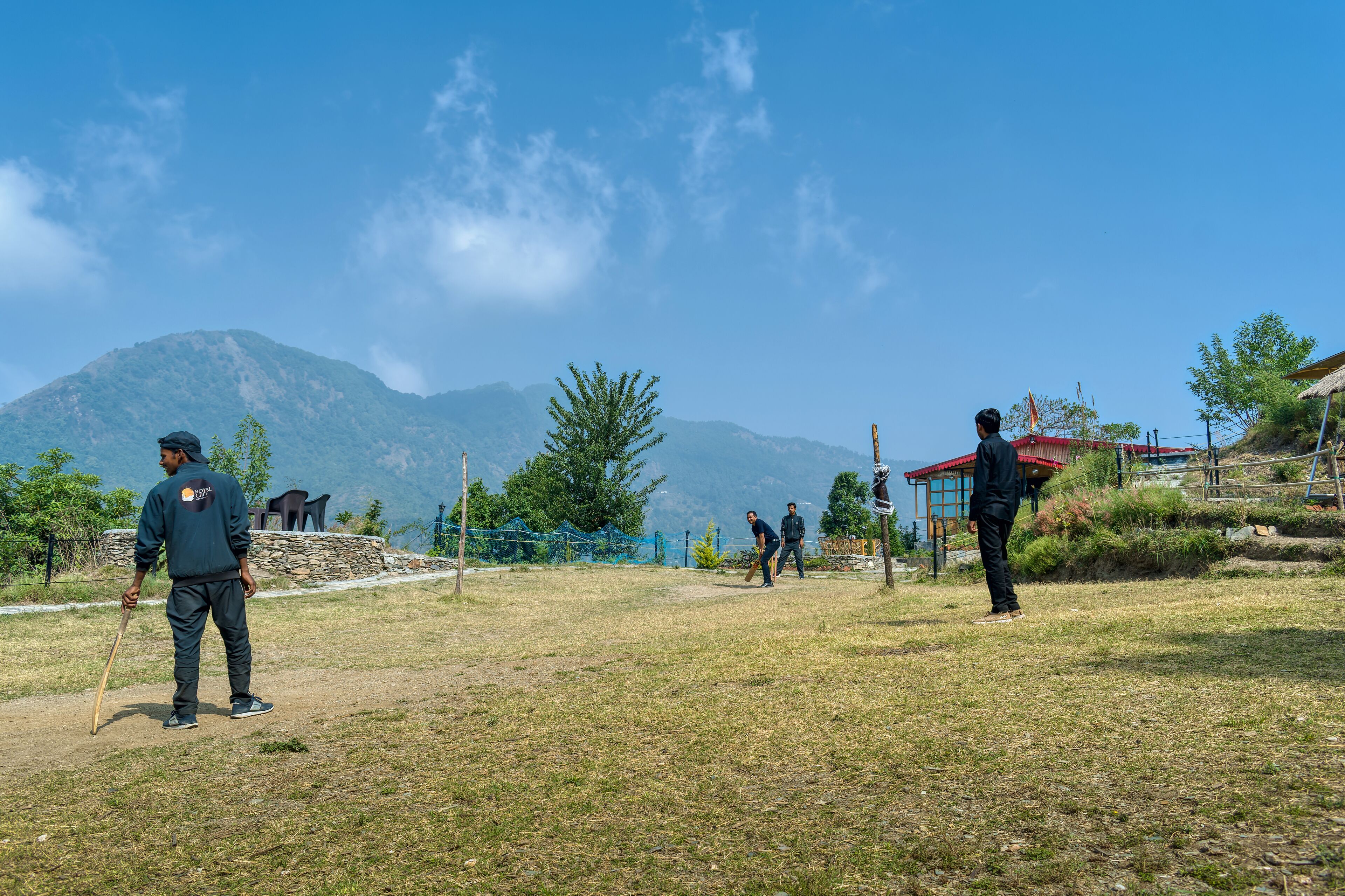 A grassy outdoor area with a mountainous backdrop, a few trees, and a building with a red roof.