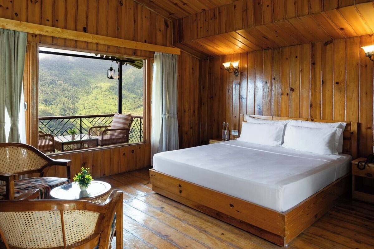 A wooden cabin room with a bed, a table with chairs, and a balcony with a view of the forest.