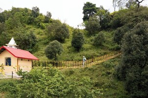 A rustic building with a red roof and a small tower, surrounded by lush greenery and a yellow suspension bridge.