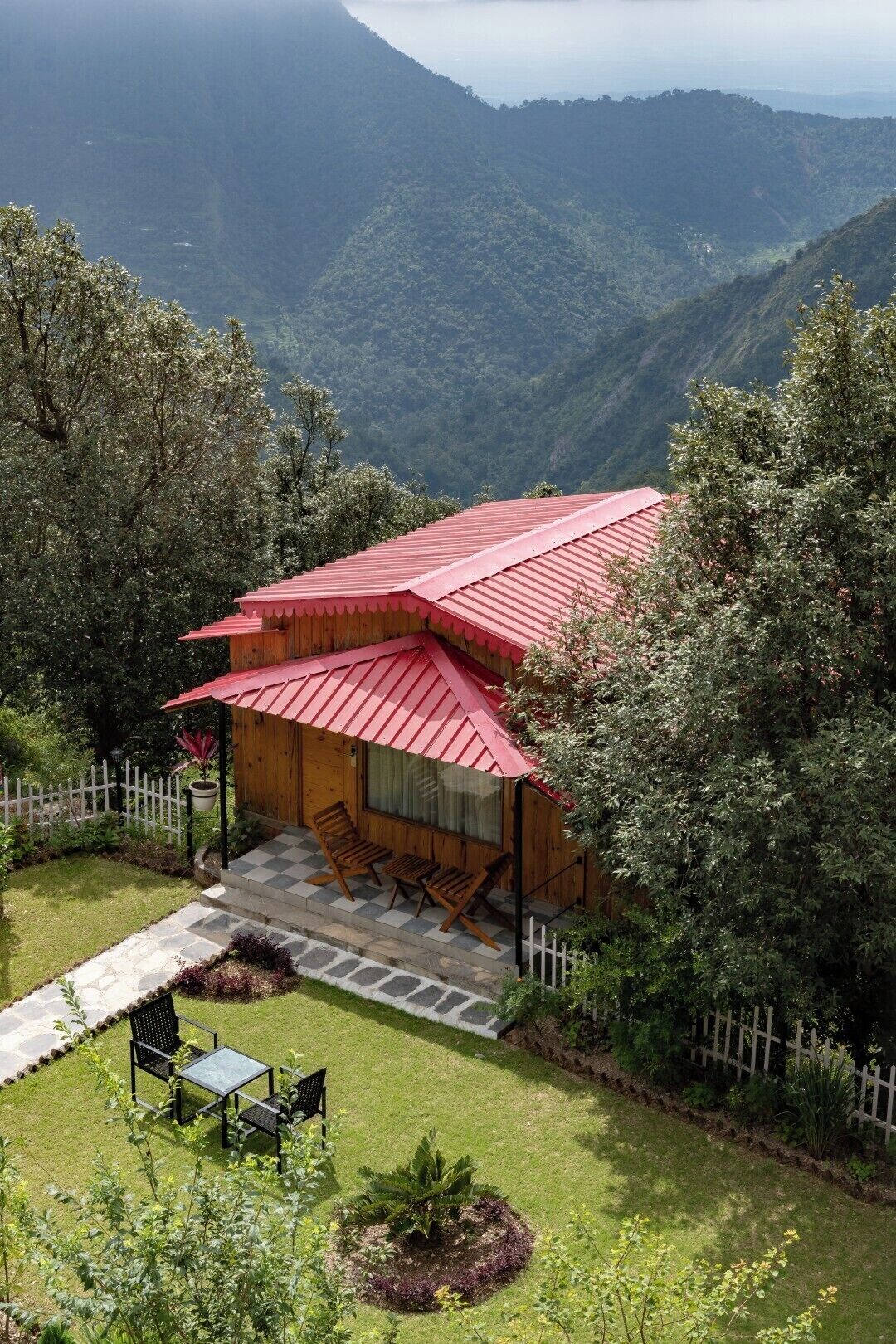 A wooden cabin with a red roof, surrounded by lush greenery and mountains in the background.