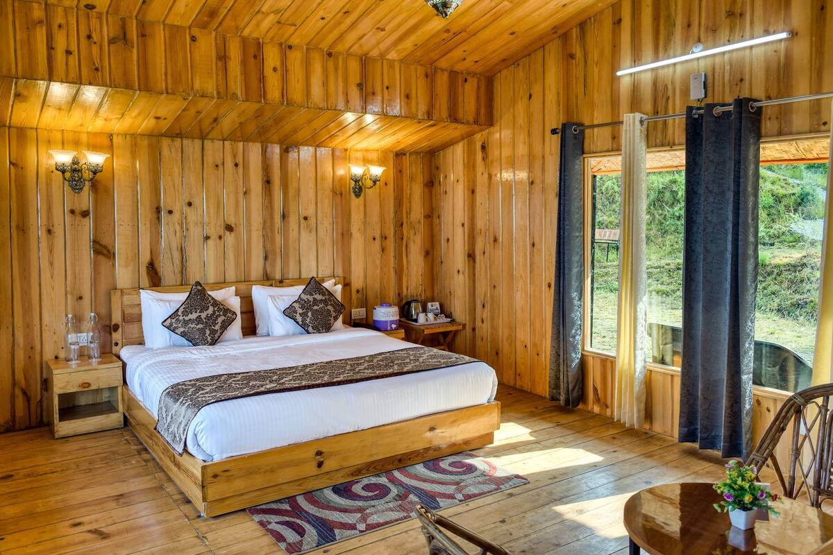 A wooden cabin bedroom with a bed, bedside tables, and a view through curtains.