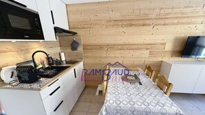 A compact kitchen with a microwave, sink, and stove, wooden wall paneling, and a dining area with a patterned tablecloth and chairs. - Studio Cabin in center Valloire with Free Wifi (VALLOIRE)