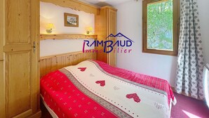 Un dormitorio con un armario de madera, una cama con colcha de rayas rojas y blancas, un pequeño estante con lámparas y una ventana con cortinas.