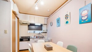 A compact kitchen with white cabinets, a sink, and a stove. There are two chairs and a table in the dining area. The walls are decorated with colorful artwork. - North Shinjuku house (Tokyo)