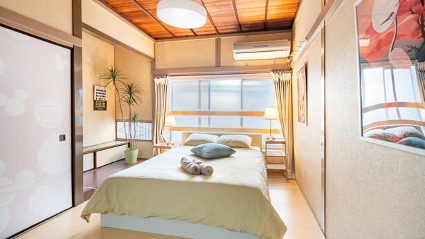 A bedroom with a bed, a bedside table, a lamp, a window, and a wall-mounted air conditioner. - North Shinjuku house (Tokyo)