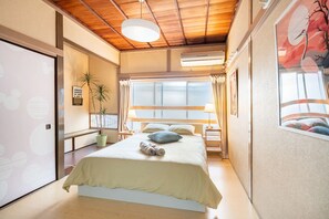 A bedroom with a bed, a bedside table, a lamp, a window, and a wall-mounted air conditioner.
