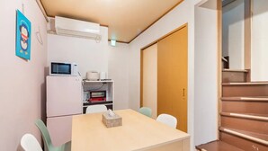 A compact kitchen with a microwave, toaster, and small refrigerator. There is a table with chairs and a box of tissues on it. A wooden door and a staircase are visible in the background. - North Shinjuku house (Tokyo)